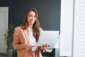 Young smiling woman with dark long hair in stylish suit working at laptop in the bright modern office photo