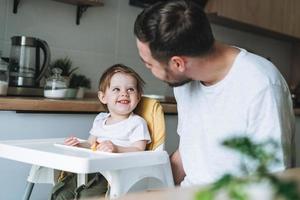 Happy father young man feeds baby girl little daughter in kitchen at home photo