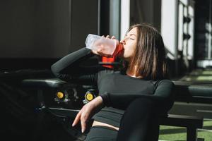 Portrait of resting young brunette woman in sport active wear drinking water in the fitness club gym photo