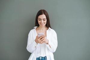 Portrait of beautiful smiling woman student with dark long hair in white shirt using mobile phone in hand on grey background isolated, generation Z photo