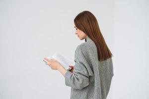 Beautiful young woman with dark long hair in grey knitted sweater reading book on the white background photo