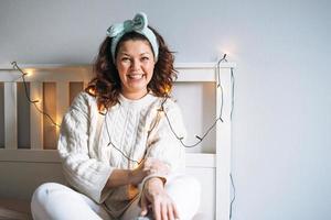 retrato de una joven morena feliz adulta de cuarenta años de cuerpo positivo en suéter de punto blanco en la cama en un hogar acogedor foto