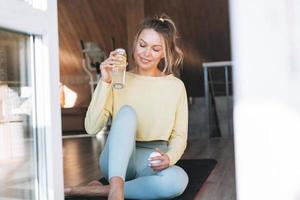 joven mujer rubia fitness con botella de agua con limón haciendo ejercicios matutinos cerca de la ventana de casa