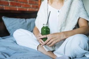 Crop photo of Young woman in casual clothes with green smoothie sitting on bed at the home