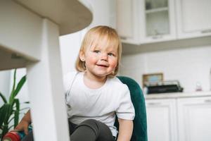 linda y graciosa niñita en silla para alimentarse en la luminosa cocina de casa foto