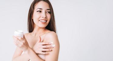 Beauty portrait of happy smiling asian woman with dark long hair put day nourishing moisturizer cream on clean fresh skin face and hands on white background, banner photo