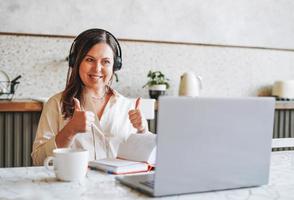 Adult smiling brunette woman in headphones doing notes in daily book with opened laptop at home photo