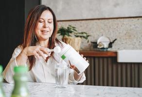 Morena sonriente adulta mujer de mediana edad cincuenta años desayunando con batido de suplemento vitamínico de proteína instantánea seca en la cocina de casa foto