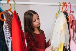 Thinking tween little girl with long dark hair in red dress among her beautiful dresses in wardrobe in the children's room at home photo