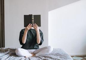 Young woman with dark long hair in casual clothes reading book on bed at the home photo