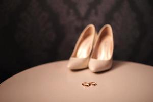 dos anillos de oro de boda y zapatos femeninos de novia de la novia. compromiso. la decoración de la boda detalla los elementos de la novia y el novio en la mesa blanca. la mañana de la novia foto