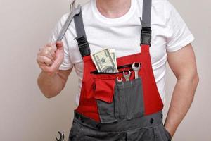 cropped photo of worker in red uniform, protective hard hat holding dollars, cash money and wrench on white background. Male worker for advertisement. world economic crisis and job loss concept