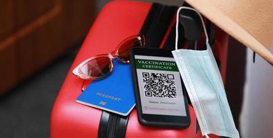 New normal concept. Close up view of a smartphone with an immune digital health passport, a red suitcase, protective mask, straw hat, passport and sunglasses. Traveling by plane during a pandemic. photo