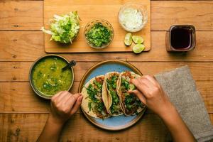 Woman's hand holding a taco of marinated meat. Plate with tacos, sauce and vegetables on wooden table. Top view. photo