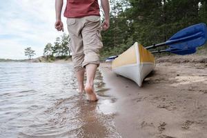 un hombre camina descalzo sobre el agua del lago hasta su kayak, que está estacionado en una playa de arena. estilo de vida. foto