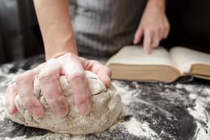 Baker holds one hand on the dough and the other on the cookbook, on the table sprinkled with flour. Bread making. Close-up. photo