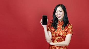 mujer china asiática con cheongsam rojo o qipao sosteniendo un teléfono móvil para la celebración del año nuevo chino aislada en el concepto de fondo rojo foto