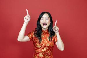 Asian woman with red cheongsam or qipao pointing to present for Chinese New Year celebration holiday isolated on red background concept photo