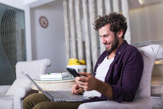 Successful entrepreneur smiling in satisfaction as he checks information on his laptop computer while working in a home office. Young man relaxing on the sofa with a laptop and mobile phone in hands photo