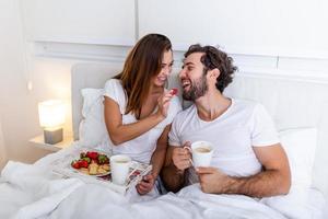 linda pareja desayunando en la cama en el dormitorio. bella mujer alimentando a su novio con fresas en la cama mientras desayuna y toma café en el dormitorio foto