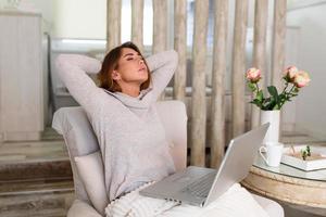 Overworked young woman sitting in her home with a laptop in front of her stretching her arms above her head with eyes closed to relax muscles, reduce tension, improve vision, photo