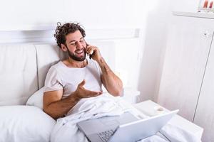 young businessman working happy and relaxed from bed with laptop computer using mobile phone in workaholic concept , Working from home in the morrning and drinking coffe photo