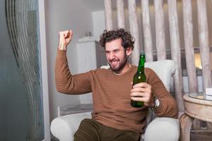 Fanatic sports fan man watching soccer game on tv celebrating. Attractive, happy guy watching football, celebrating victory of his favorite team, having beer , sitting in living room photo