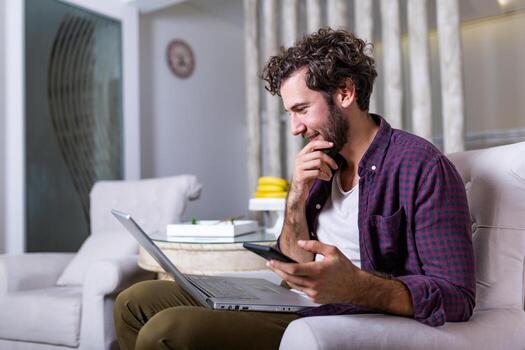 Successful entrepreneur smiling in satisfaction as he checks information on his laptop computer while working in a home office. Young man relaxing on the sofa with a laptop and mobile phone in hands photo