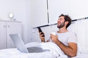 young businessman working happy and relaxed from bed with laptop computer using mobile phone in workaholic concept , Working from home in the morrning and drinking coffe photo