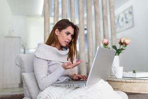 atractiva mujer loca sentada en su sofá en casa, mirando la pantalla de un portátil con expresión enojada. demasiada carga de trabajo, error de archivo, todos los datos fallaron, olvidé guardar el documento foto