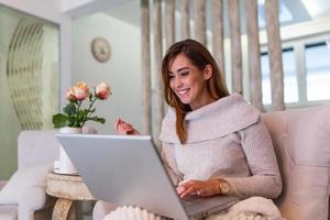 Positive young woman freelancer watching video in blog on netbook and having a coffee at comfortable home. Beautiful young business woman working on laptop.Freelancer connecting to internet photo