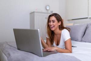 Smiling woman lying down the bed in front of her laptop with her legs raised. Happy casual beautiful woman working on a laptop sitting on the bed in the house. Freelance working from home concept photo