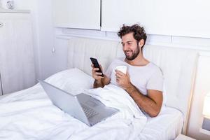 young businessman working happy and relaxed from bed with laptop computer using mobile phone in workaholic concept , Working from home in the morrning and drinking coffe