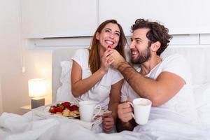 pareja enamorada desayunando en la cama. joven pareja caucásica desayunando romántico en la cama. hembra y macho, dos tazas de café, frutas y galletas coloridas. foto