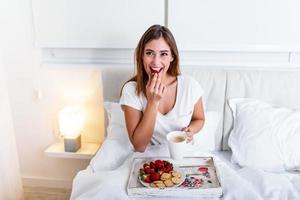 Breakfast in bed for young beautiful woman. Woman having coffee and breakfast in bed with fruits, coffee and biscuits. Morning waking up in hotel photo