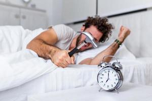 Young man tries to break the alarm clock with hammer, Destroy the Clock. Man lying in bed turning off an alarm clock with hammer in the morning at 7am. photo