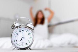 reloj despertador de primer plano que tiene un buen día con antecedentes mujer feliz estirándose en la cama después de despertarse, luz del sol por la mañana. despertarse por la mañana a tiempo foto