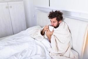 hombre enfermo con gripe acostado en la cama controlando su temperatura y bebiendo bebidas calientes. hombre enfermo acostado en la cama con fiebre alta. gripe fría y migraña. foto