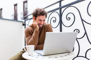 Happy young man smiling, as he works on his laptop to get all his business done early in the morning with his cup of coffee photo