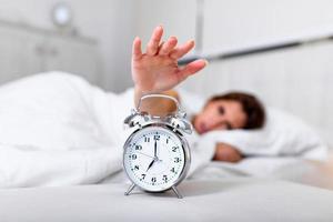 Woman lying in bed turning off an alarm clock in the morning at 7am. Hand turns off the alarm clock waking up at morning, girl turns off the alarm clock waking up in the morning from a call. photo