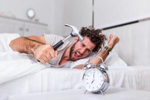 el joven intenta romper el despertador con un martillo, destruir el reloj. hombre acostado en la cama apagando un despertador con un martillo por la mañana a las 7 am. foto