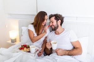 linda pareja desayunando en la cama en el dormitorio. bella mujer alimentando a su novio con fresas en la cama mientras desayuna y toma café en el dormitorio foto