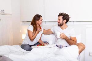 Cute couple having breakfast in bed in the bedroom. Beautiful woman feeding her boyfriend strawberries in bed while having breakfast and coffee in bedroom photo