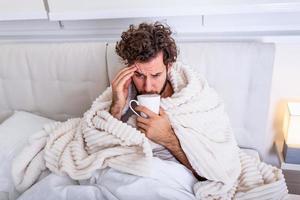 Portrait of a sick man with the flu, allergy, germs,cold coughing. Sick Man with headache sitting under the blanket with high fever and a flu, resting and drinking hot beverage photo
