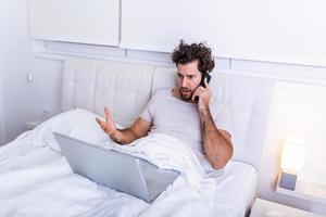 Image of young Caucasian man has morning coffee, sits on bed, working on his laptop and talking on his mobile phone, prepares for examination, uses laptop computer for surfing internet. photo