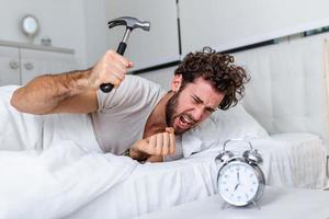 el joven intenta romper el despertador con un martillo, destruir el reloj. hombre acostado en la cama apagando un despertador con un martillo por la mañana a las 7 am. foto