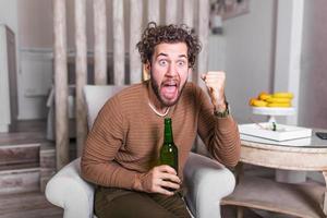 joven emocionado animando mientras ve un partido deportivo en el sofá en casa. chico atractivo y feliz viendo fútbol, celebrando la victoria de su equipo favorito, gritando, tomando cerveza. foto