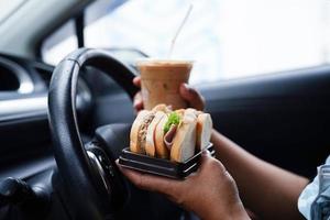 Asian woman driver hold ice coffee cup and sandwich bread for eat and drink in car, dangerous and risk an accident. photo