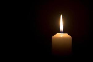 una sola llama de vela encendida o luz que brilla en una vela blanca sobre fondo negro u oscuro en la mesa de la iglesia para Navidad, funeral o servicio conmemorativo foto
