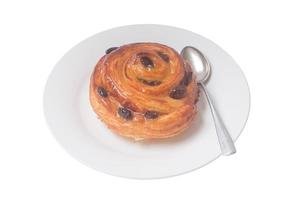 pastel danés en espiral o bollo dulce con pasas encima en un plato blanco con una cucharadita pequeña servida para el desayuno aislado en fondo blanco foto